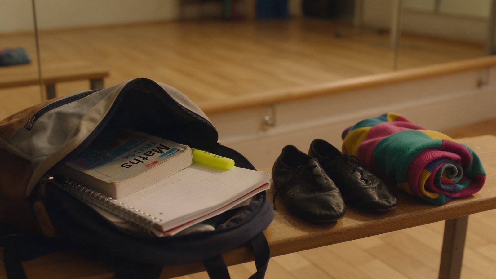 Una motxilla d'institut oberta amb llibres i apunts, al costat d'unes sabatilles de dansa, sobre el banc d'una sala de dansa, amb la barra i el mirall al fons