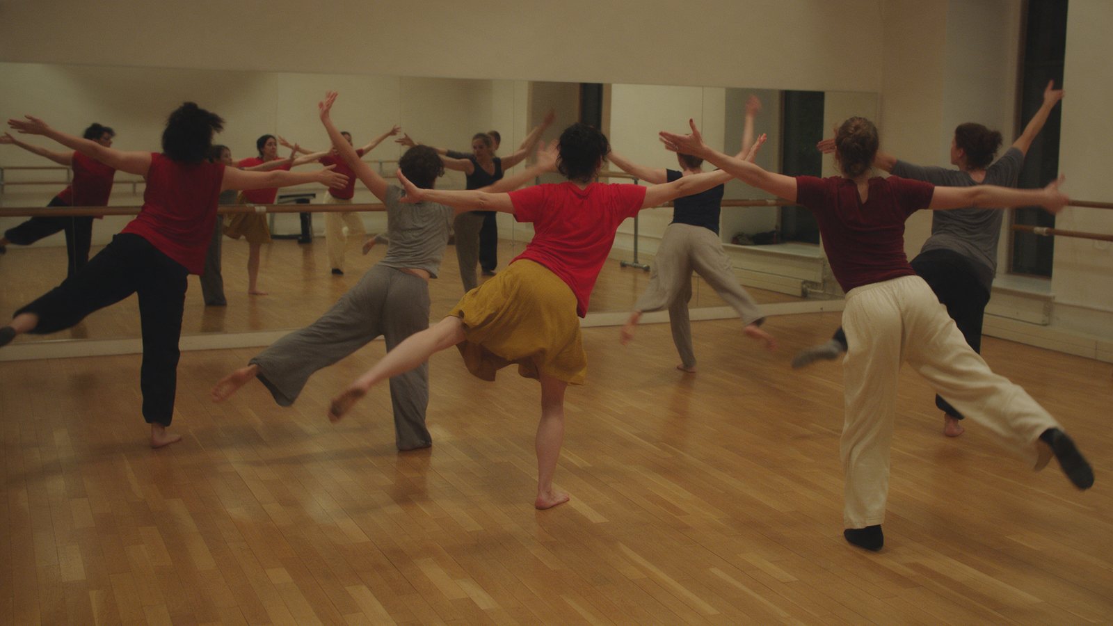 Un grupo de alumnas de espaldas, a medio movimiento con una pierna atrás y los brazos abiertos, ante el espejo y la barra de una sala de danza